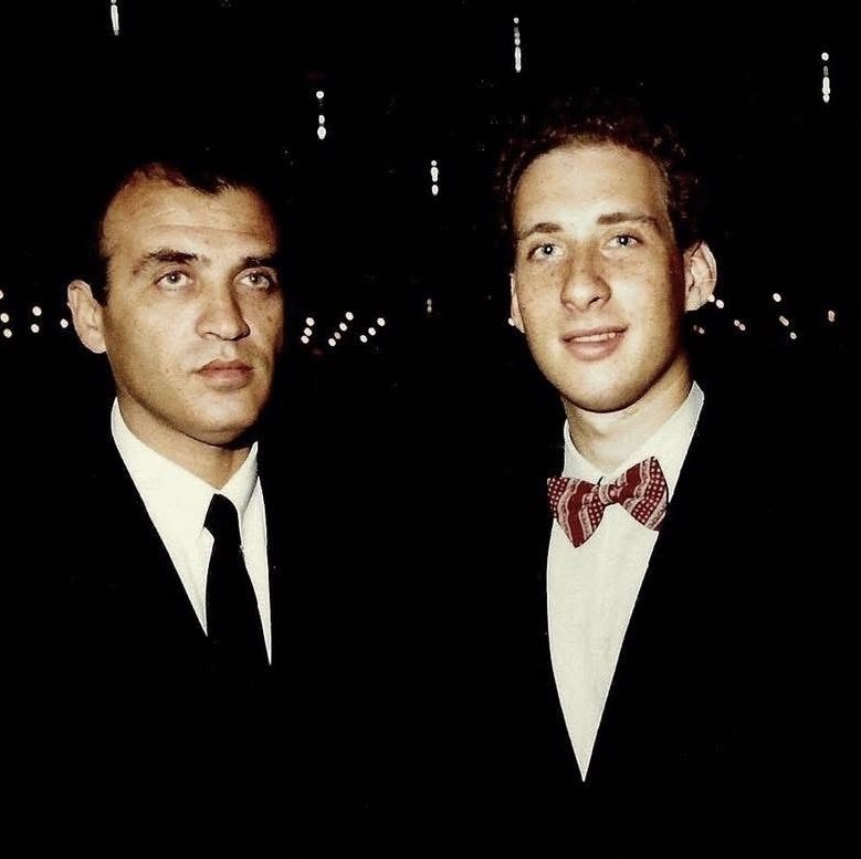 Two men in black tie at an evening reception, lights behind them