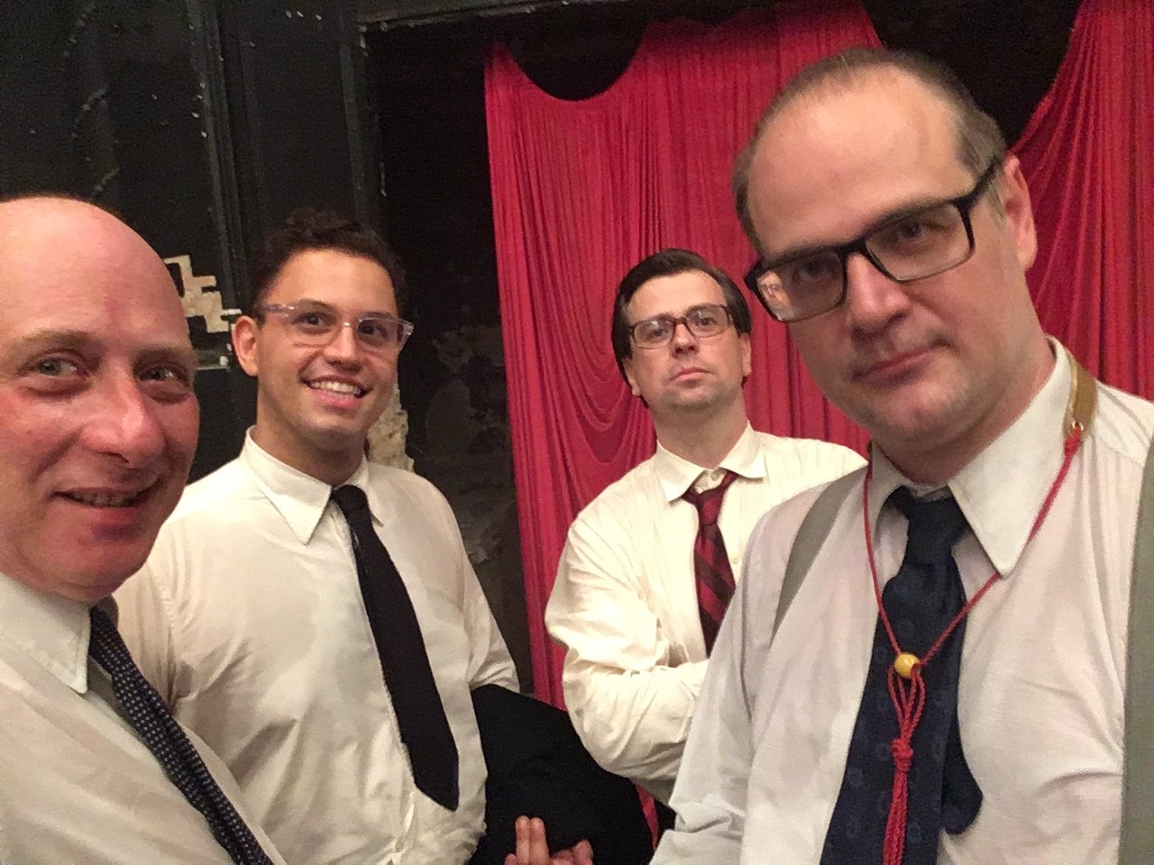 Four musicians in period shirts and ties backstage before a red curtain