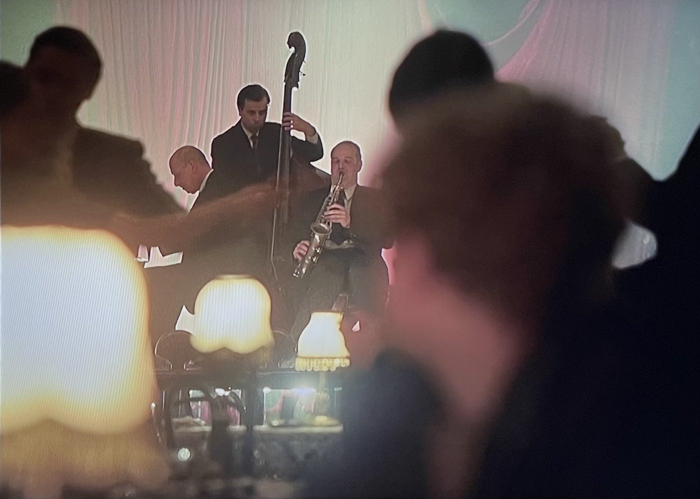 A jazz trio playing in a club scene seen past the silhouettes of seated guests