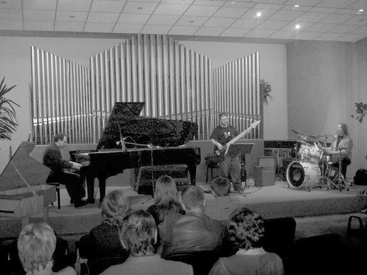 The trio on a concert platform before tall organ pipes, piano at the left, bass in the centre, drums at the right