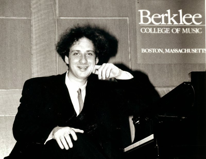 Grego with curly hair leaning on a grand piano and smiling, a Berklee College of Music sign on the wall behind