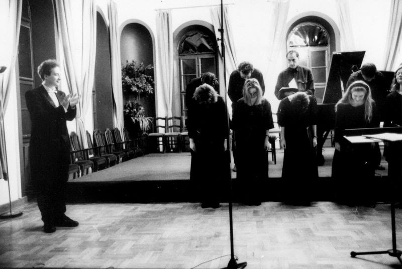 Grego conducting Cantus Corvinus in a hall with tall curtained windows, the singers bowing at the end of the piece
