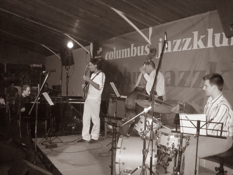 A quartet on stage under a Columbus Jazzklub banner, Grego at the piano on the left
