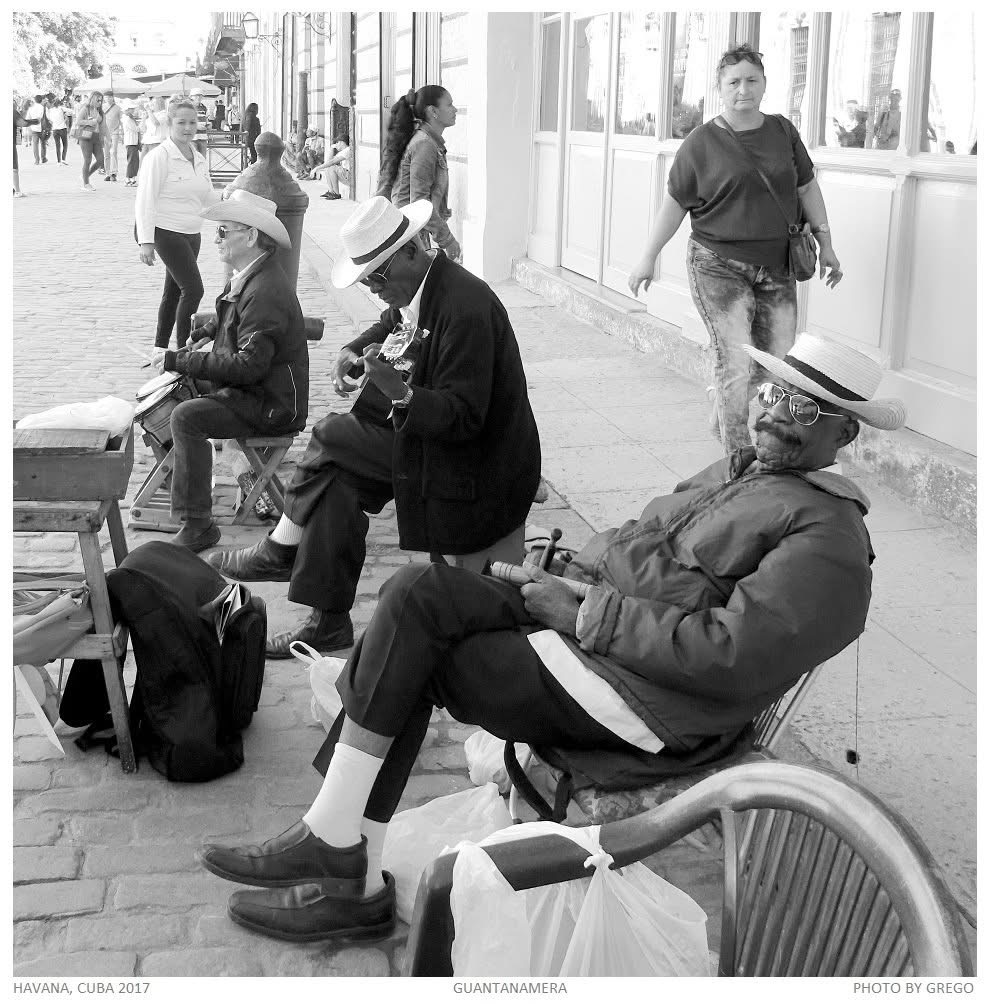 Three street musicians in straw hats resting on a Havana pavement as passers-by walk past