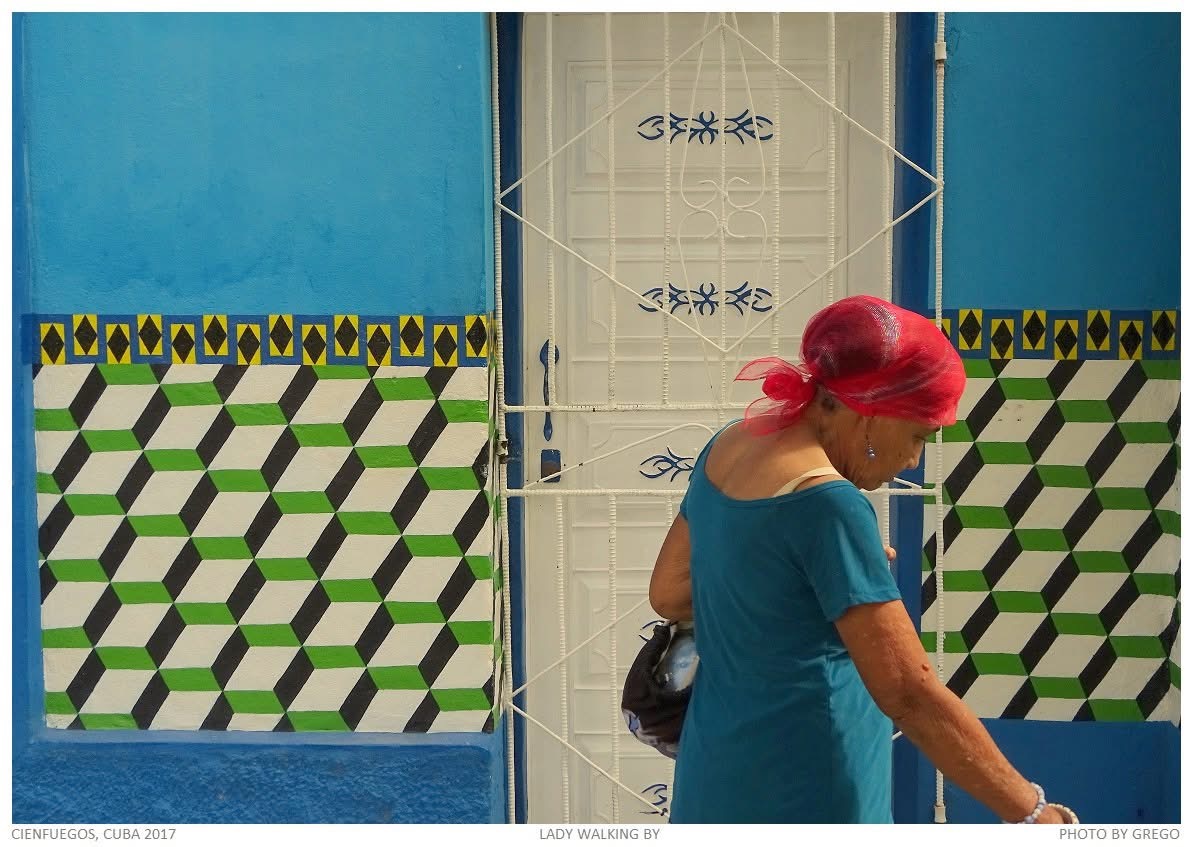 A woman in a red headscarf and teal dress passing a blue wall painted with green cube patterns