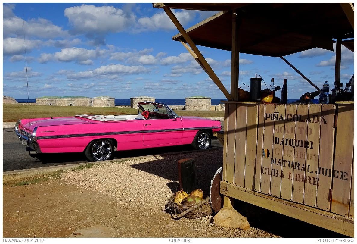 A pink convertible parked beside a wooden drinks stand listing piña colada, mojito and Cuba libre