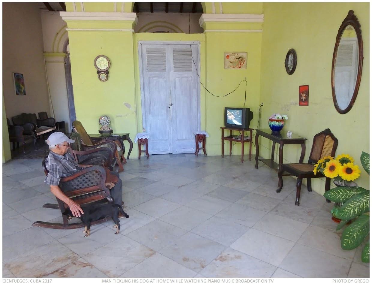 An elderly man in a rocking chair tickling his dog in a bare green room while piano music plays on a small television