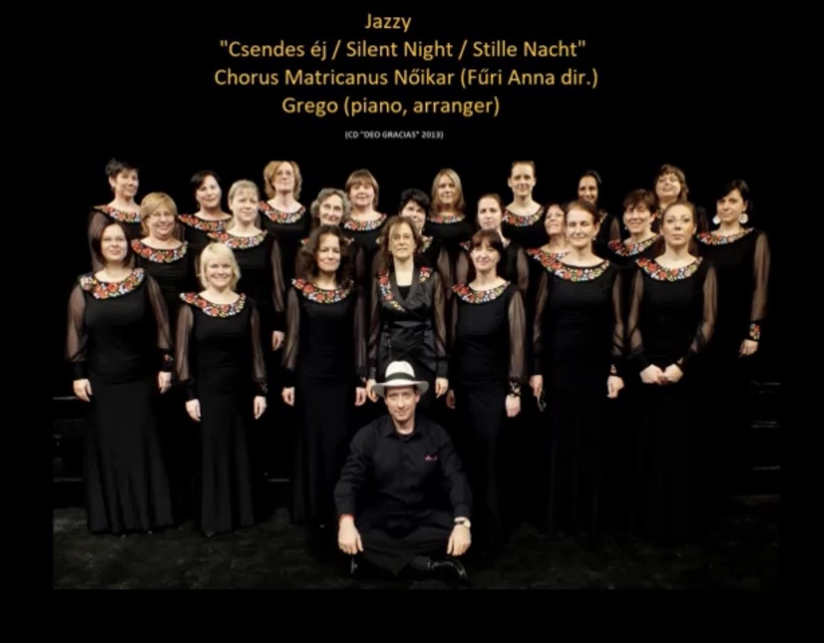Chorus Matricanus on stage in black dresses with embroidered collars, Grego seated in front in a white hat
