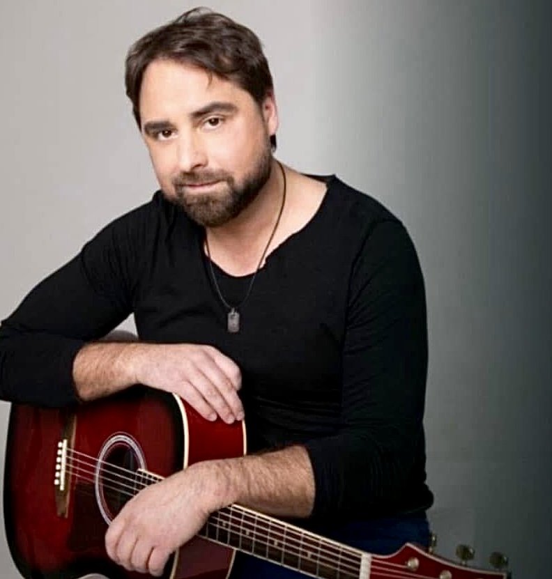 Gyula Éliás Jr. seated with an acoustic guitar, against a plain grey background