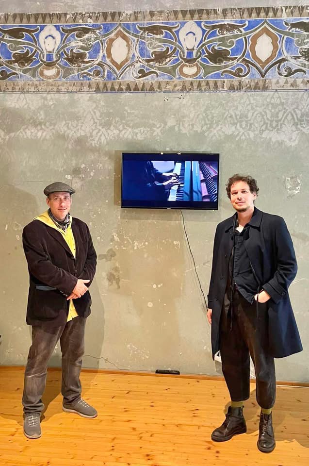 Ladislav Pálmai and a colleague beside a wall-mounted screen showing hands at a keyboard, in a gallery with exposed frescoes
