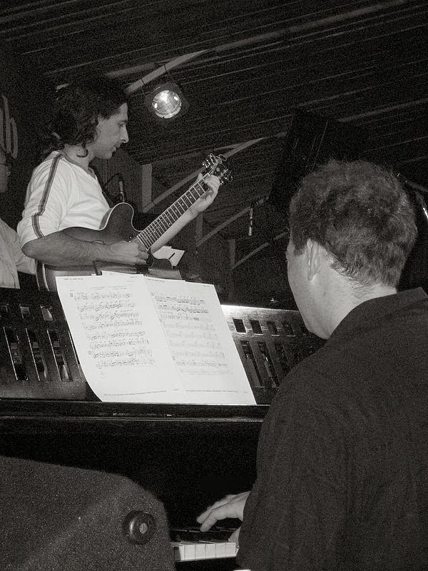A guitarist playing beside Grego at the piano in a club, a mirror ball above