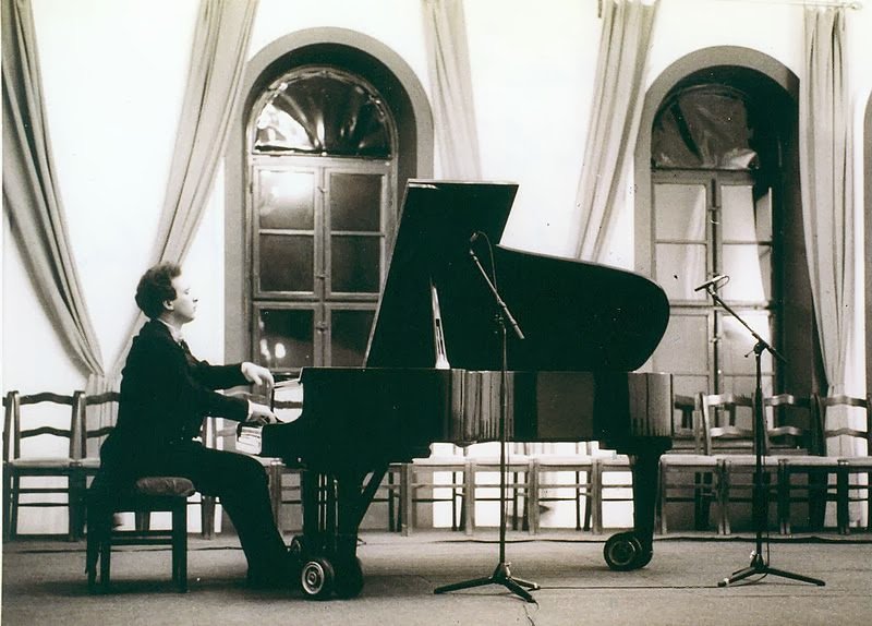 Grego alone at a grand piano on a small stage with tall curtained windows