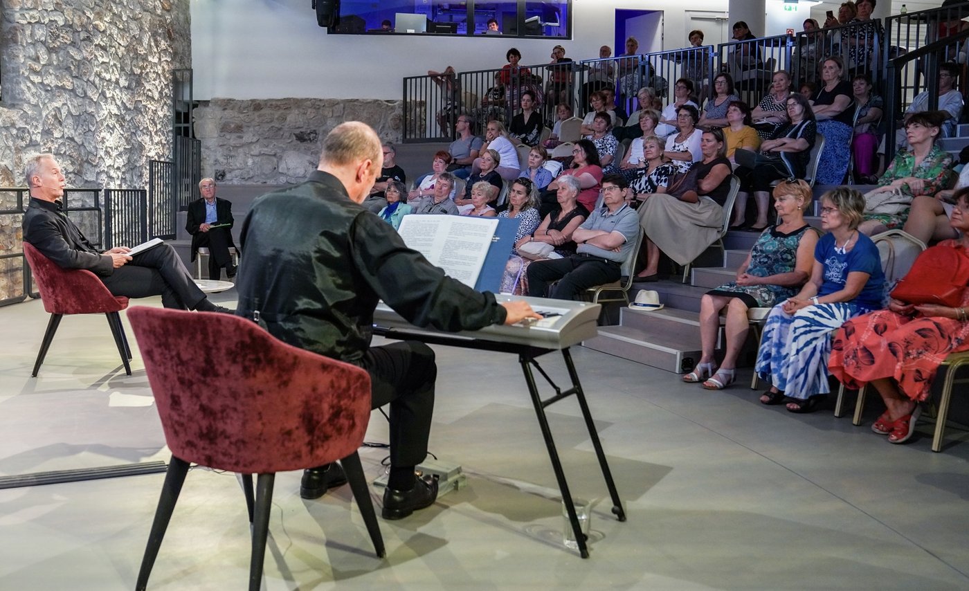 A reader and Grego at a keyboard on a stone-walled stage, the audience banked on steps opposite