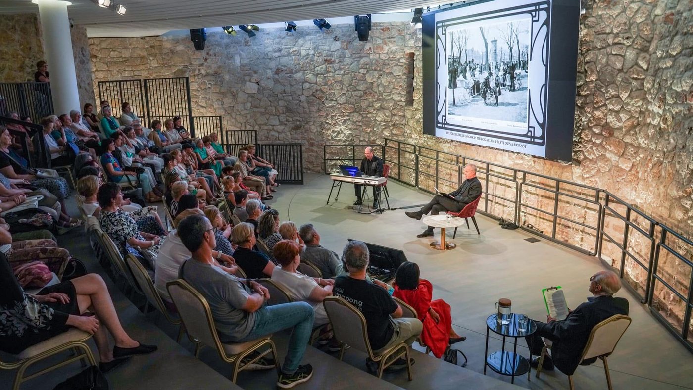 Grego at a keyboard beside a reader on a stone-walled stage, an old Budapest photograph projected behind