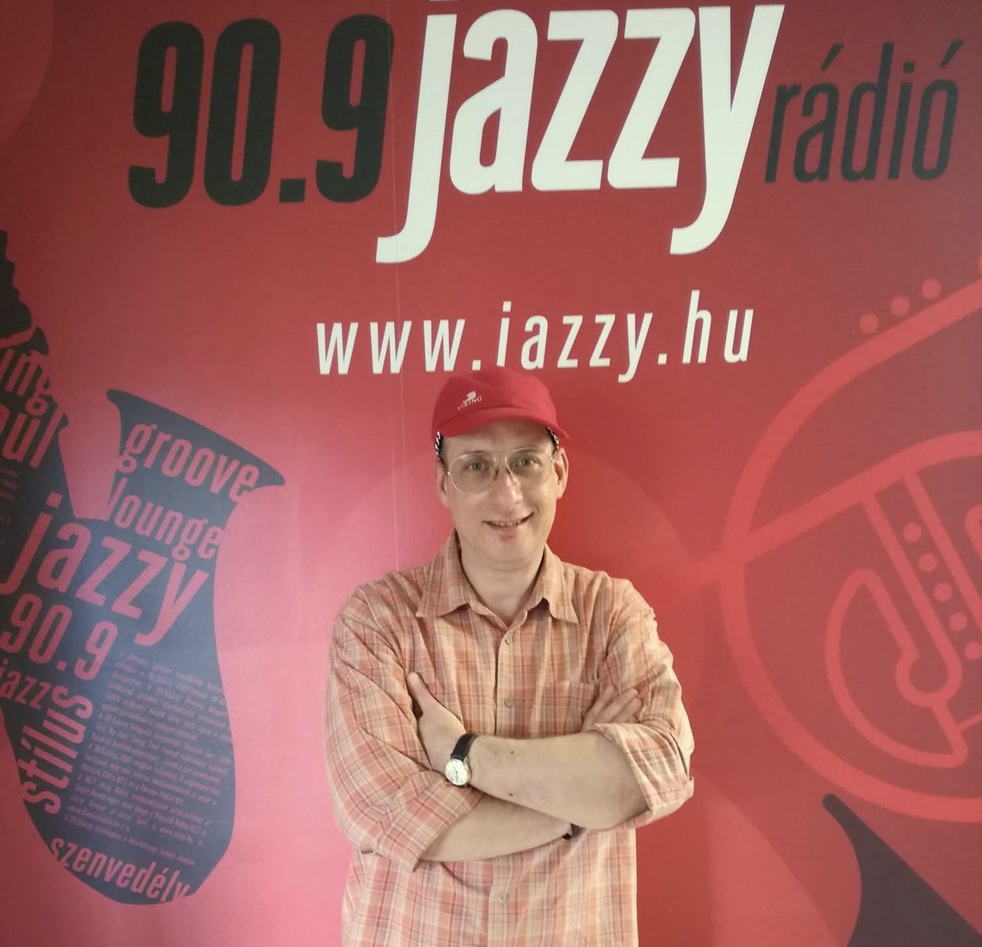 Grego standing with folded arms in front of a large Jazzy Rádió 90.9 backdrop