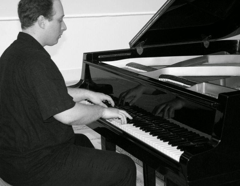 Close view of Grego at the keyboard of a black grand piano, his hands reflected in the lid