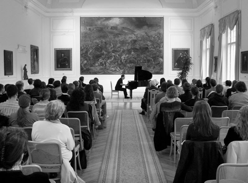 Grego alone at a grand piano at the far end of a panelled hall beneath a large battle painting, the audience seated in rows