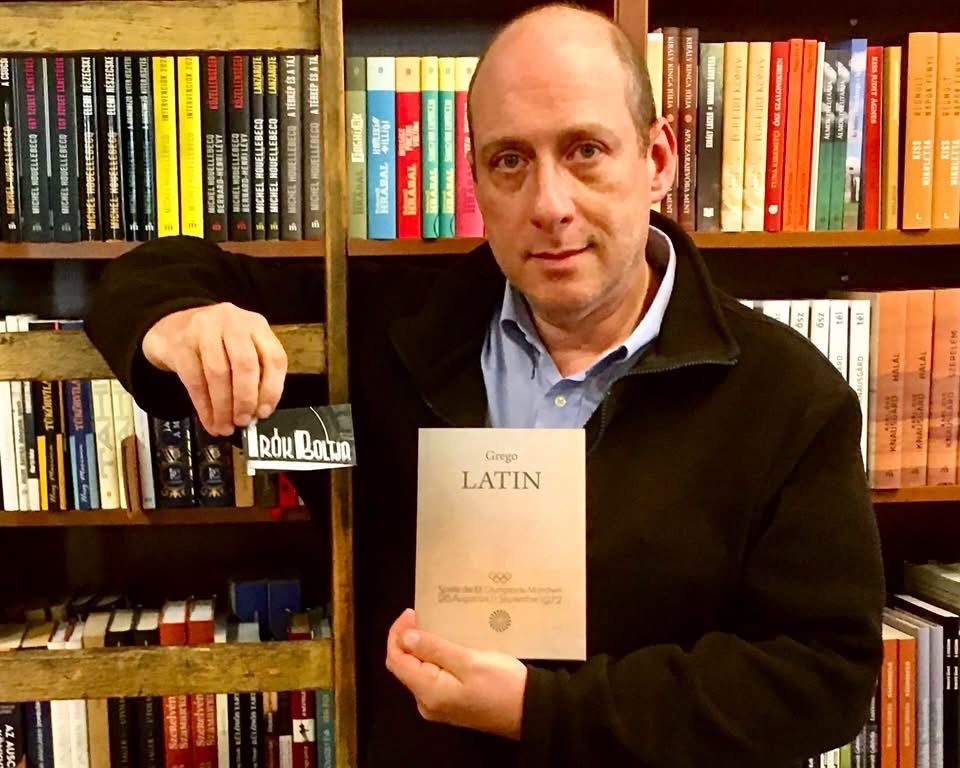 Grego holding a copy of Latin in front of bookshelves in a bookshop