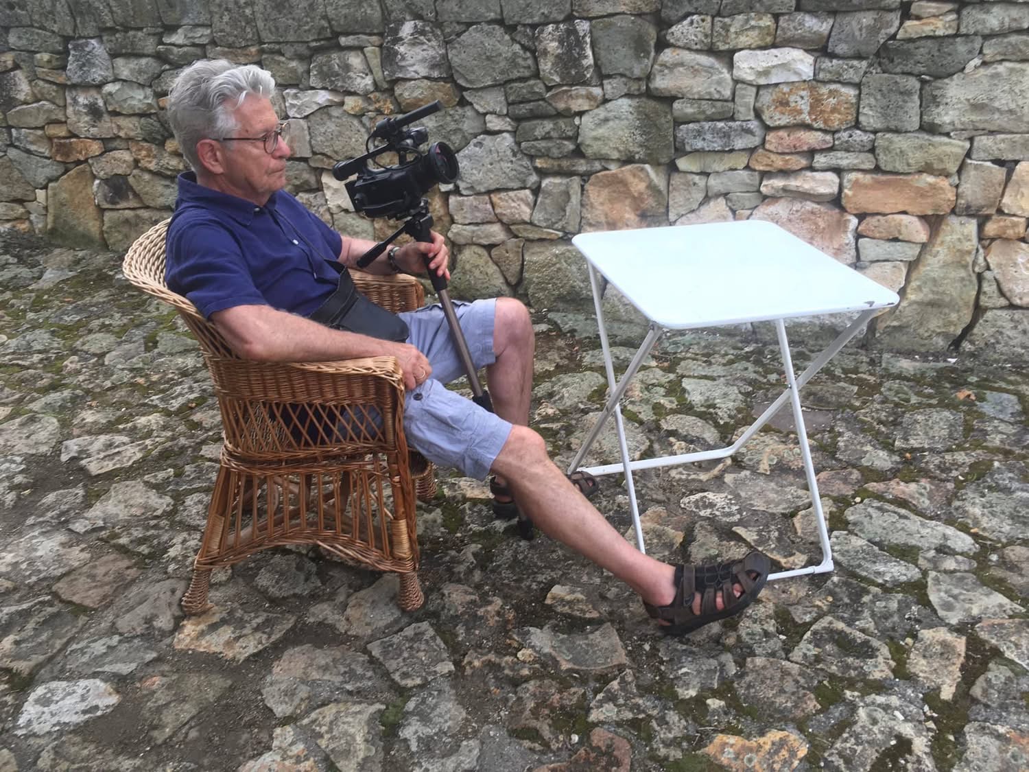 István Jávor seated in a wicker chair filming beside a stone wall