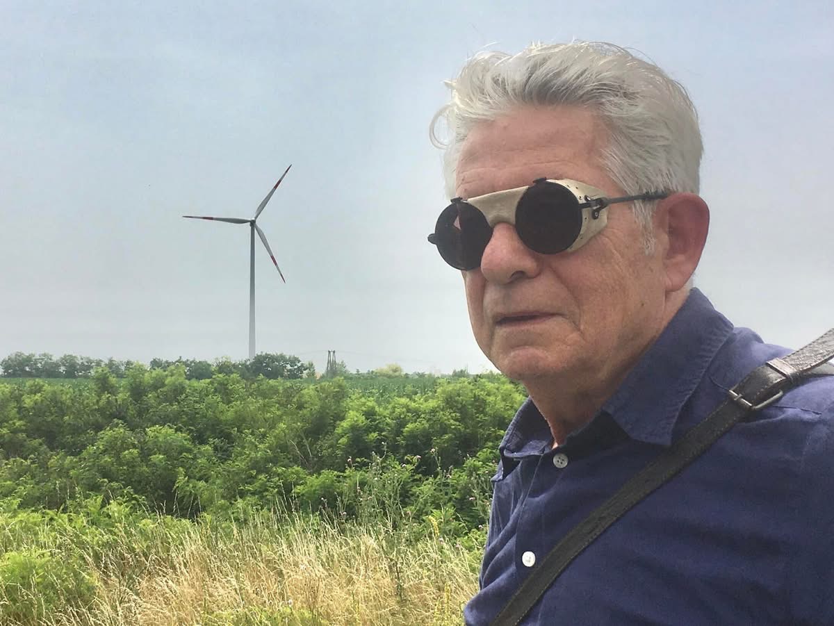 István Jávor in dark round glasses on location, a wind turbine behind him