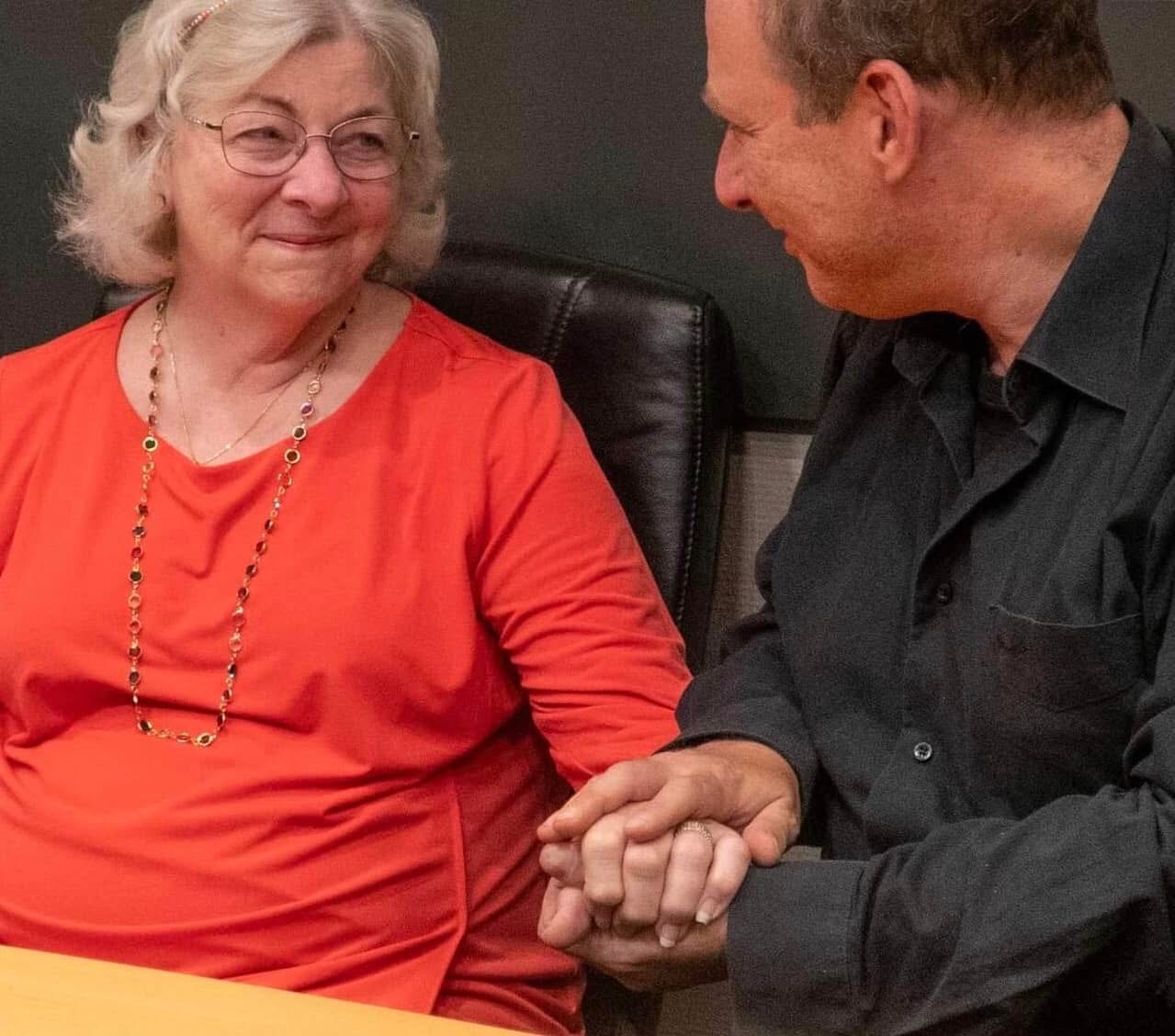Grego holding the hand of an older woman, both seated, talking