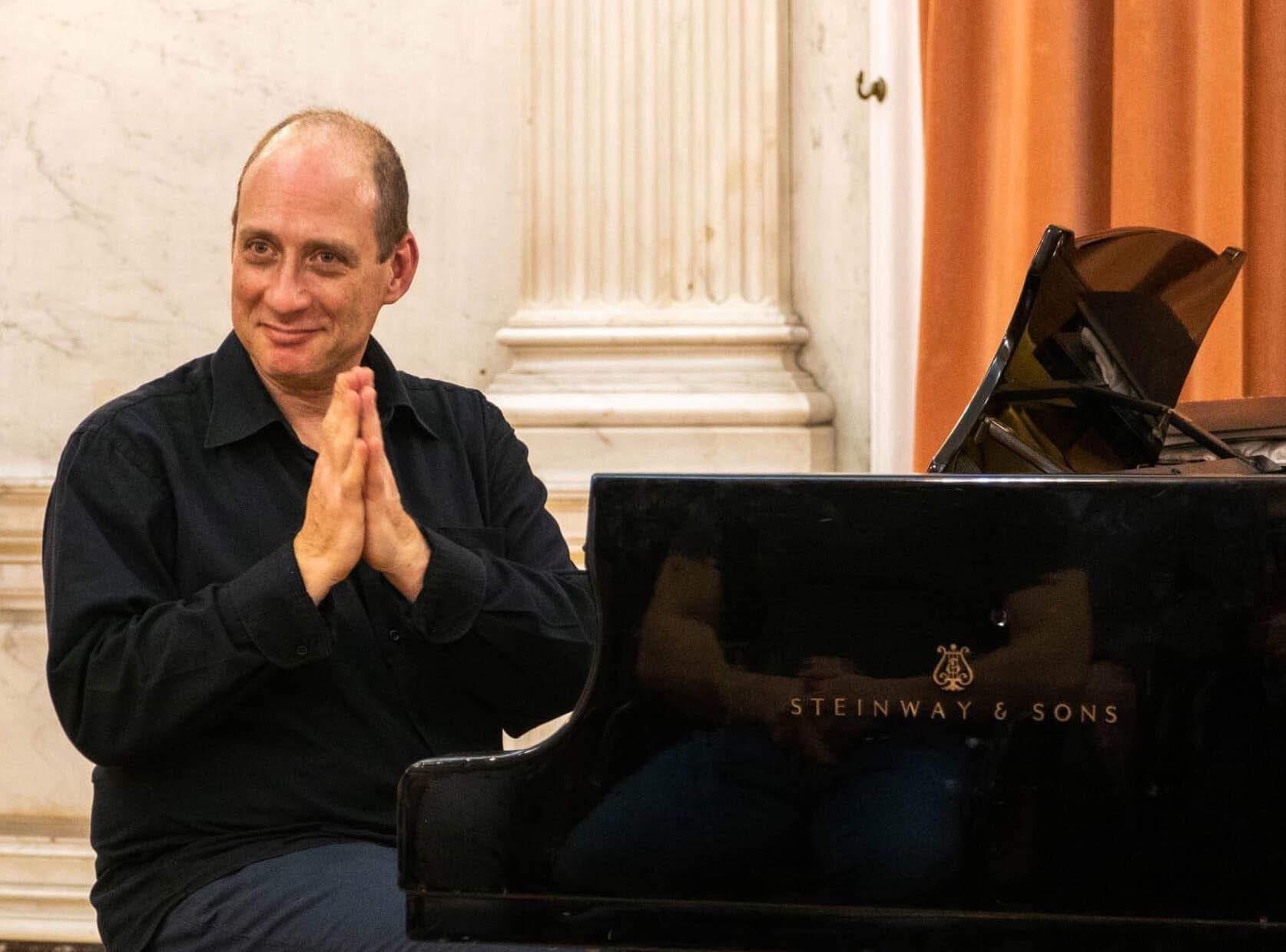 Grego seated at the Steinway with his hands pressed together, smiling
