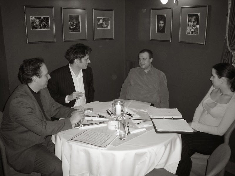 Four musicians talking around a table with scores and framed photographs on the wall behind