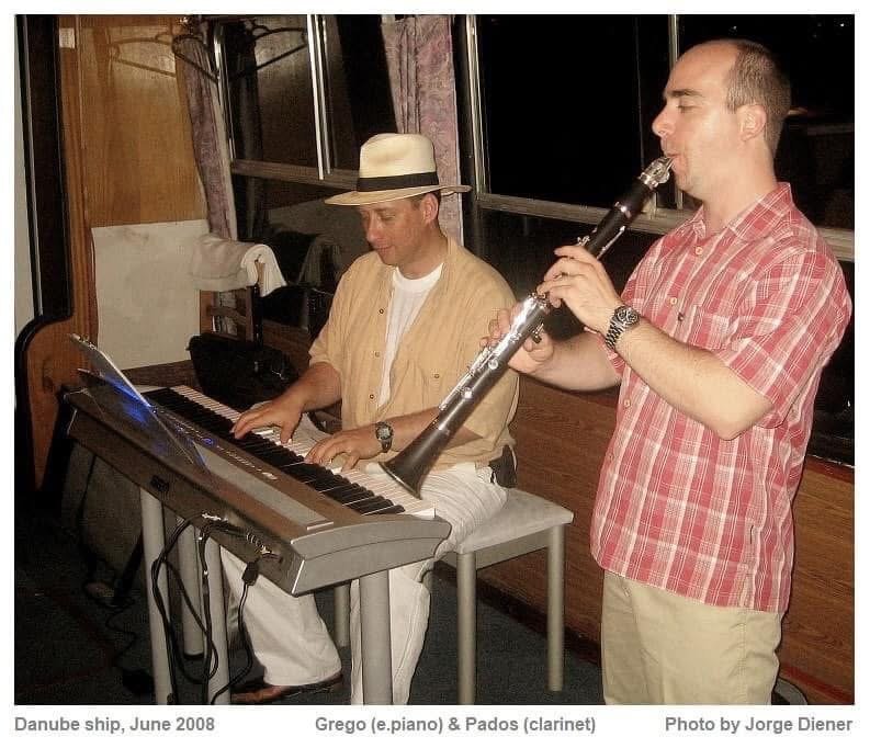 Grego at an electric piano in a white hat with a clarinettist standing beside him on a boat