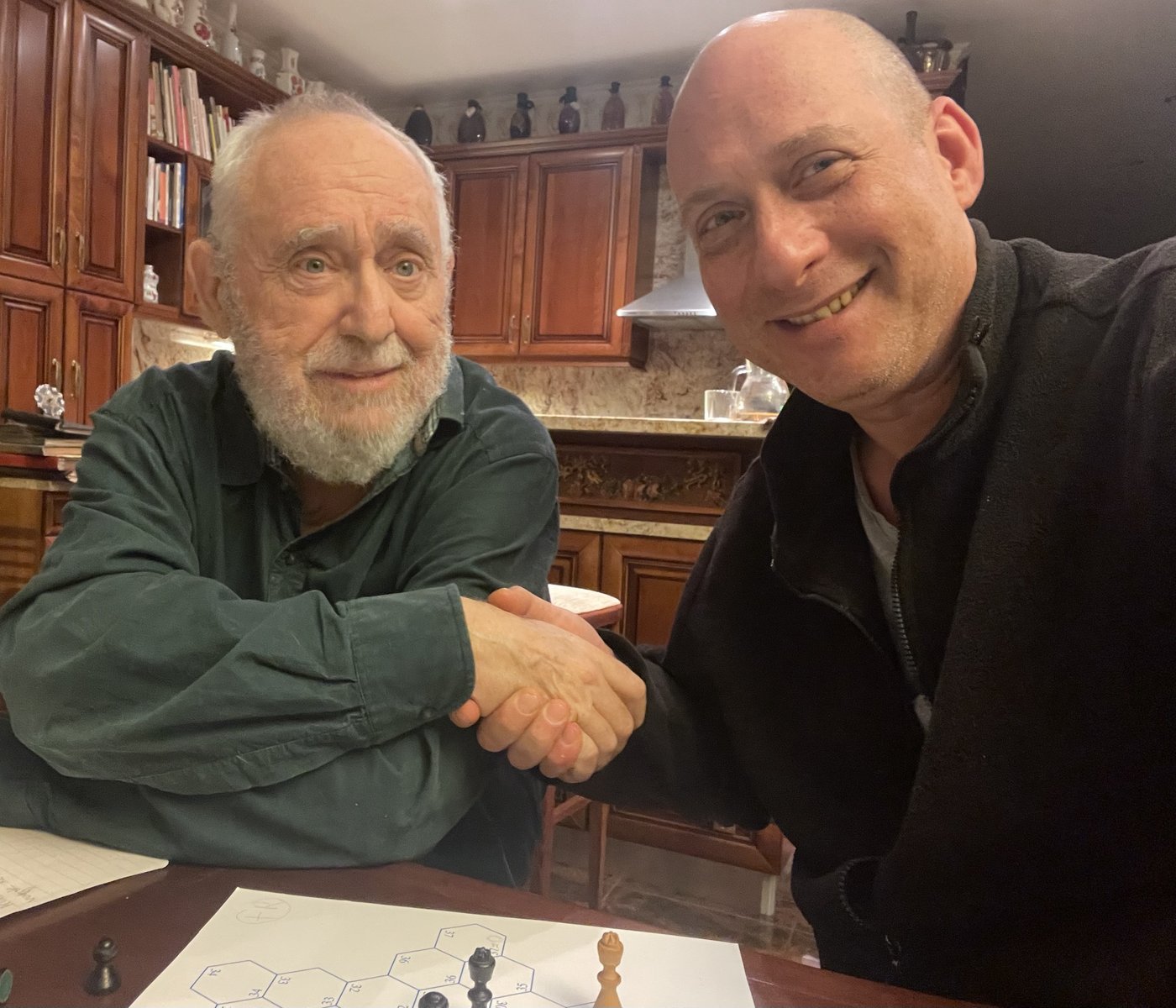 Grego shaking hands with László Polgár across a table, a hexagonal chess diagram and pieces in front of them