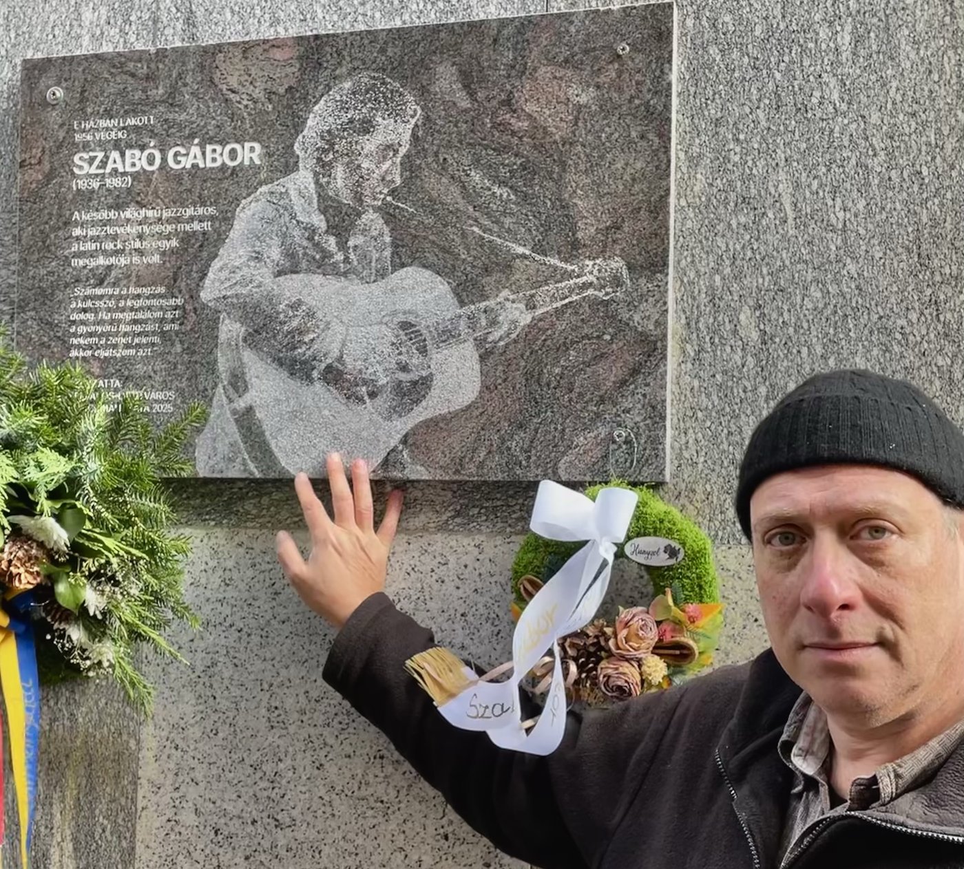 Grego with his hand on a granite memorial plaque to Gábor Szabó, wreaths below it