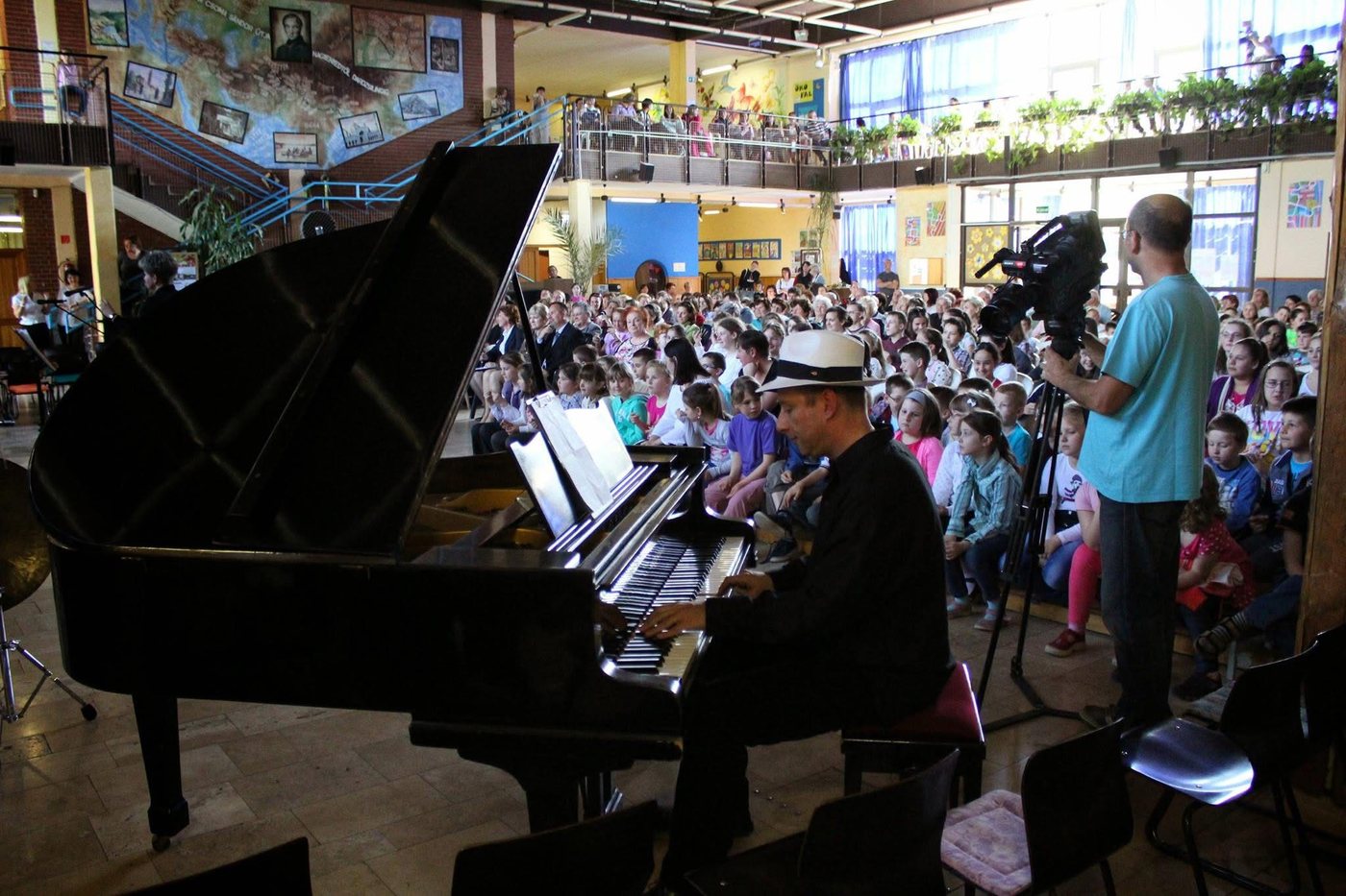 Grego in a white hat playing a grand piano to a school hall packed with children