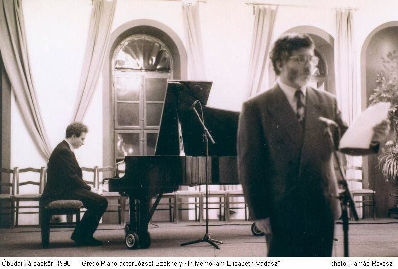 Grego at the grand piano at the back of the hall while an actor reads at a microphone in the foreground