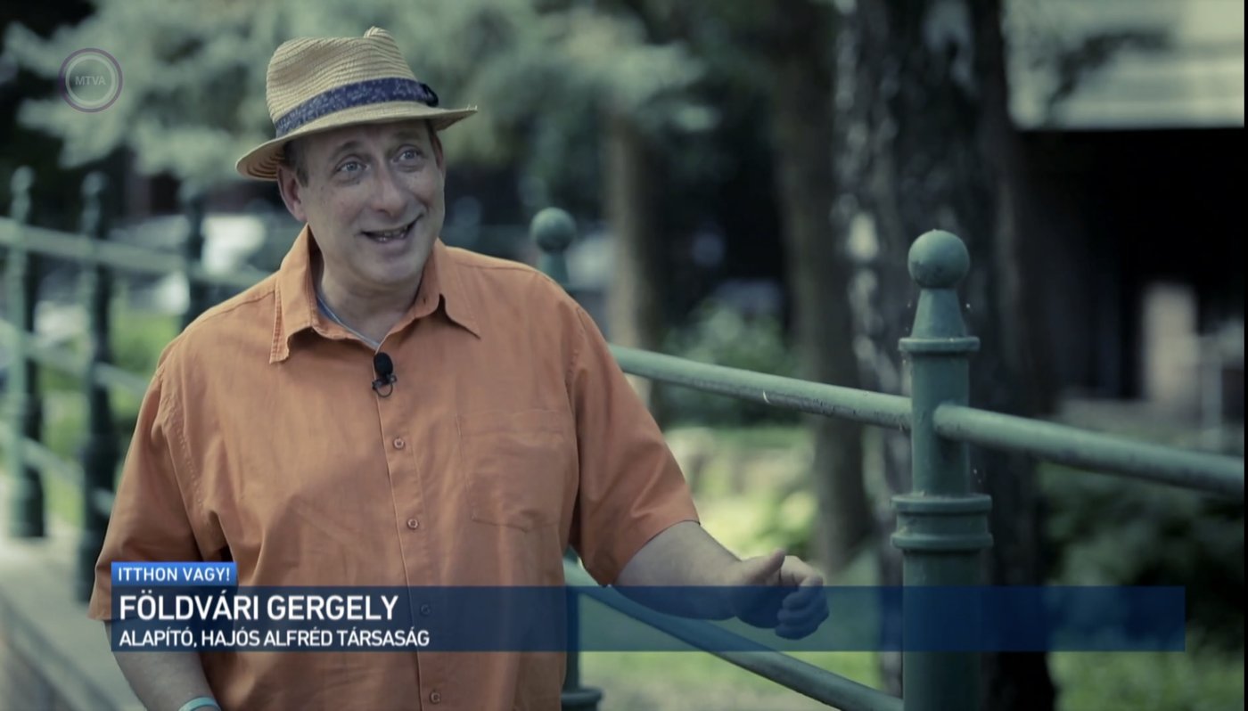 Grego in a straw hat speaking to camera on a park bridge, a television caption across the foot