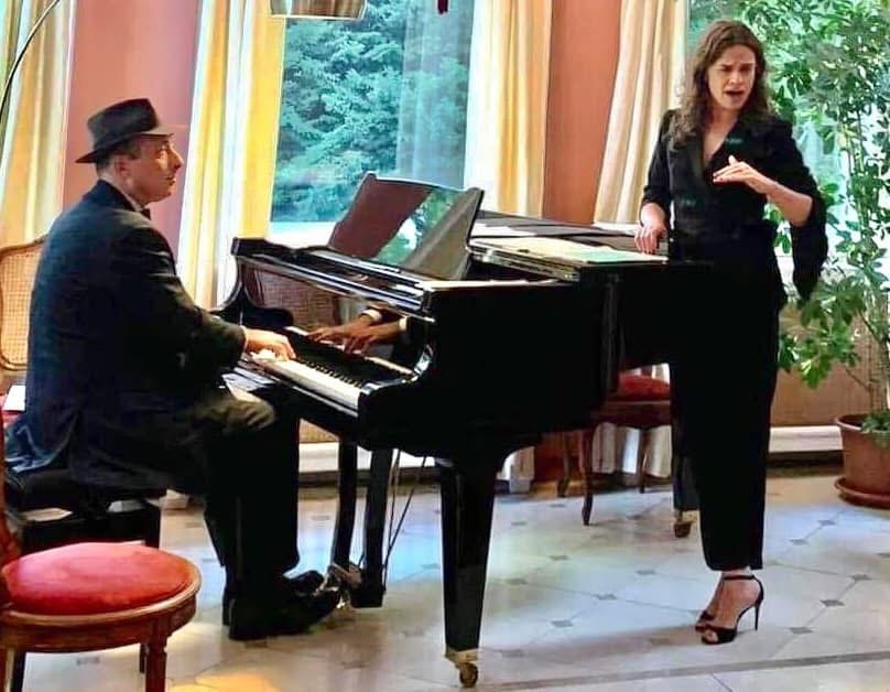 Grego at a grand piano in a reception room while a singer stands beside it