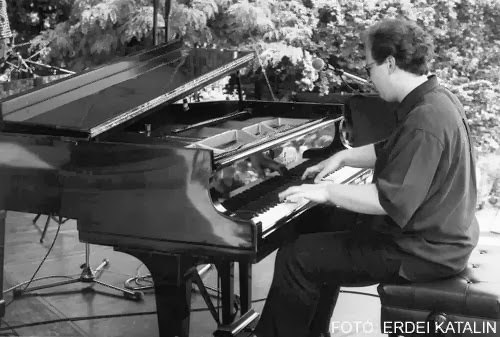 Grego at a grand piano on an outdoor stage, microphones and conifers behind him