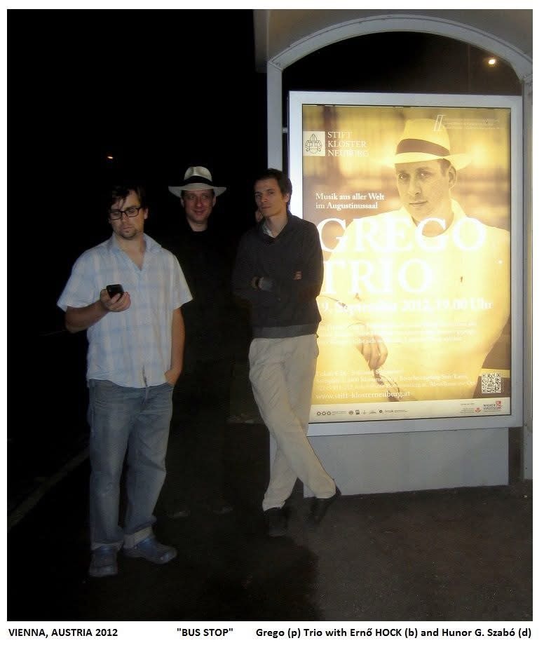 Three musicians beside a lit poster for the Grego Trio at a bus stop at night