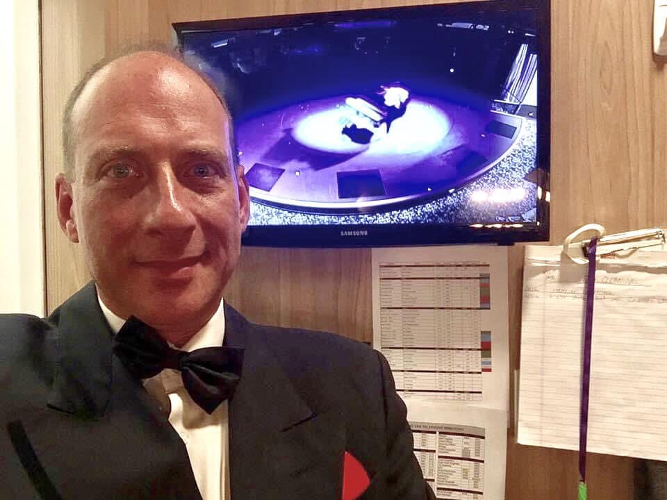 Grego in black tie backstage, the stage visible on the monitor behind him