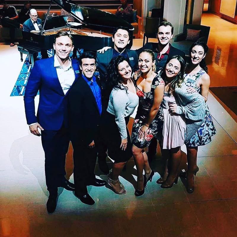 The ship’s singers and dancers gathered together, Grego at the grand piano behind them