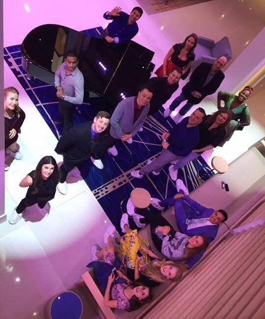 The cast looking up at the camera from around a grand piano, seen from above